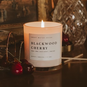 May include: A lit candle in a frosted glass jar with a gold base. The label reads "BLACKWOOD CHERRY" with scent notes. Fresh cherries are in the foreground. The candle is 11 oz.