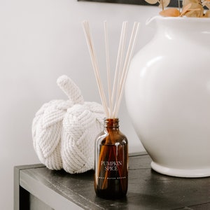 May include: A brown glass reed diffuser bottle with a label reading PUMPKIN SPICE and SWEET WATER DECOR, containing several light-colored wooden reeds. The bottle is positioned next to a white woven rope pumpkin and a large white ceramic pitcher.