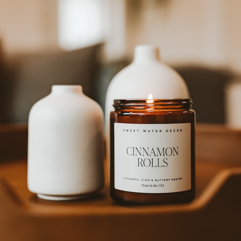 May include: A lit amber glass candle with a white label sits on a wooden surface between two white ceramic vases. The label reads 'Sweet Water Decor,' 'Cinnamon Rolls,' 'Cinnamon, Icing & Buttery Pastry,' and 'Made in the USA.' The candle and vases are home decor items with a minimalist aesthetic.