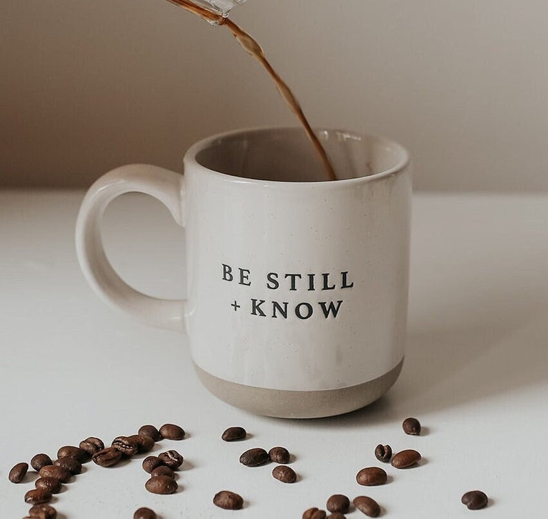 May include: A white ceramic mug with the words "BE STILL + KNOW" in black lettering is being filled with coffee. Coffee beans are scattered around the mug. The mug has a beige base and a curved handle.