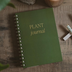 May include: A dark green spiral-bound journal with the words PLANT journal printed in gold lettering on the front cover. The notebook features a metallic wire binding along the left side and a smooth, matte finish on the solid green cover.