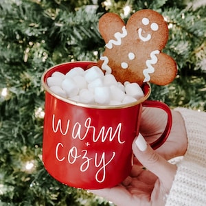 A red campfire style mug is shown with a white script "Warm and Cozy" saying on two sides and gold rim on top.