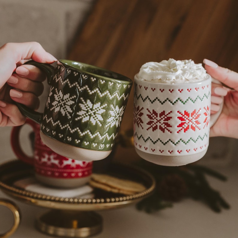 May include: Two ceramic mugs with embossed Nordic-style snowflake patterns. One mug is dark green with white patterns, and the other is white with red and green patterns. The white mug is filled with whipped cream, and both have a light-colored base.