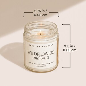 May include: A clear glass candle jar with a white candle inside. The jar is labelled "Sweet Water Decor Wildflowers and Salt" with the scent description "Jasmine, Rosemary, Sea Salt & Honey". The candle is 8.89 cm tall and 6.98 cm wide.