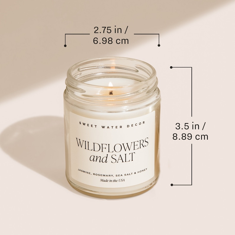 May include: A clear glass candle jar with a white candle inside. The jar is labelled "Sweet Water Decor Wildflowers and Salt" with the scent description "Jasmine, Rosemary, Sea Salt & Honey". The candle is 8.89 cm tall and 6.98 cm wide.