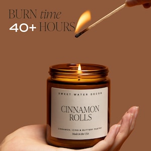 Peut inclure: Un bocal en verre brun contenant une bougie avec une &eacute;tiquette blanche sur laquelle est inscrit "SWEET WATER DECOR CINNAMON ROLLS CINNAMON, ICING & BUTTERY PASTRY Made in the USA". La bougie est allum&eacute;e et la flamme est visible.