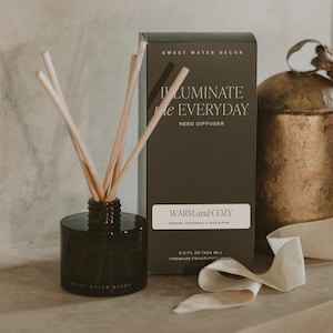 May include: A dark glass reed diffuser bottle with light wooden reeds and a matching dark green box labeled SWEET WATER DECOR ILLUMINATE the EVERYDAY WARM and COZY ORANGE, CINNAMON, CLOVE & PINE 3.5 FL OZ (104 ML) PREMIUM FRAGRANCE.