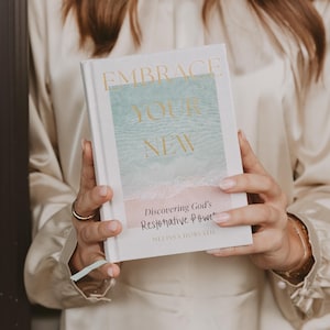 May include: A white book titled "EMBRACE YOUR NEW" with gold lettering. The cover features a watercolor design of a beach scene in shades of blue, pink, and white. The book's subtitle reads "Discovering God's Restorative Power" by Melissa Horvath.