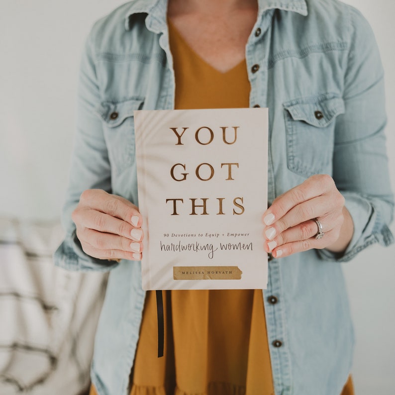 May include: A closed book with a white cover and gold lettering. The title is "You Got This" and the subtitle is "90 Devotions to Equip & Empower Hardworking Women". The author's name is "Melissa Horvath" and is printed in a gold banner at the bottom of the cover.