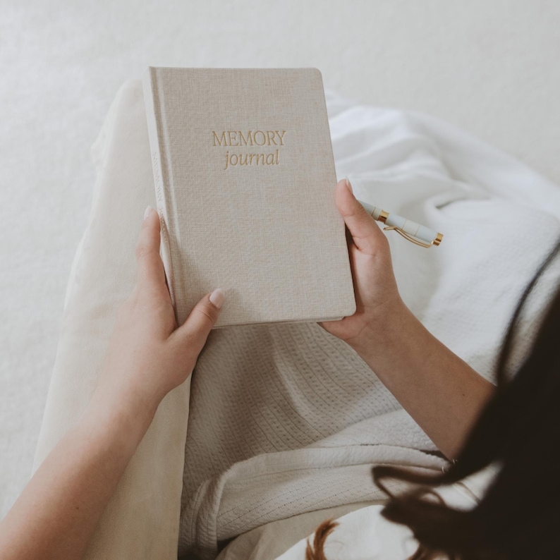 Pu&ograve; includere: Una persona tiene in mano un diario beige con le parole "MEMORY journal" stampate in oro sulla copertina.