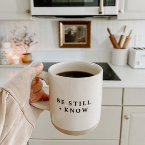 May include: A white ceramic mug filled with dark coffee, featuring the text "BE STILL + KNOW" in black. The mug has a tan base and is held in a kitchen setting. The image evokes a sense of calm and comfort.