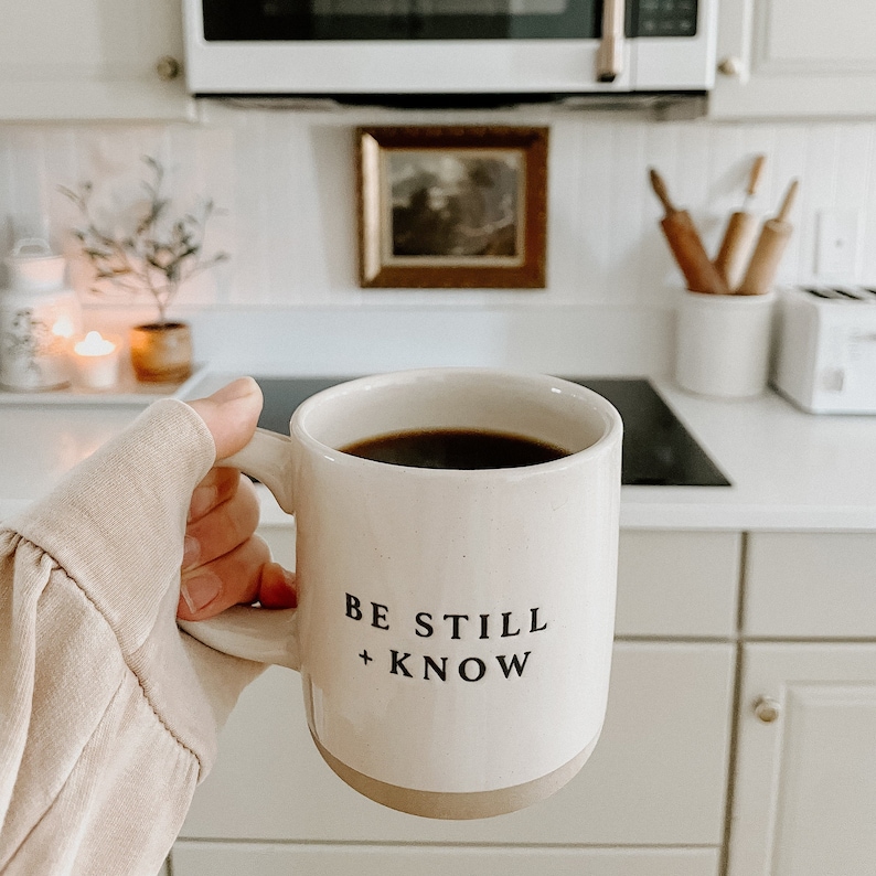 May include: A white ceramic mug filled with dark coffee, featuring the text "BE STILL + KNOW" in black. The mug has a tan base and is held in a kitchen setting. The image evokes a sense of calm and comfort.