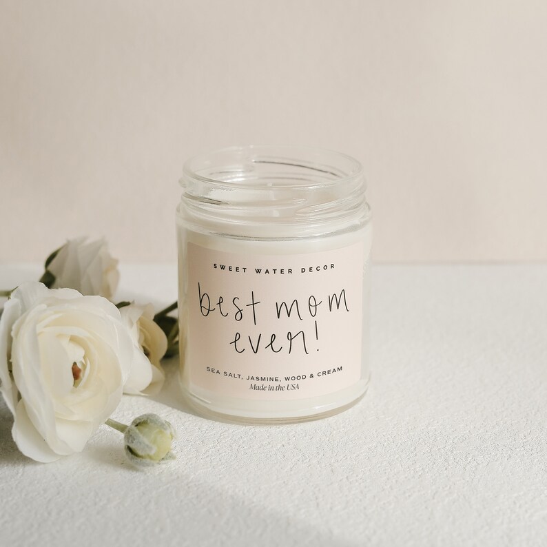 May include: A white candle in a glass jar with a white label that reads "SWEET WATER DECOR best mom ever! SEA SALT, JASMINE, WOOD & CREAM Made in the USA"