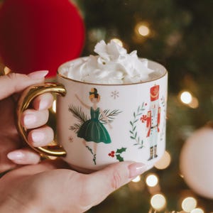 May include: A white ceramic mug with a gold handle, filled with whipped cream. The mug features illustrations of a ballerina and a nutcracker. Festive holiday decor is visible in the background.