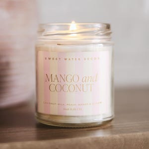 Może przedstawiać: Zapalonego świeca w szklanym słoiku z etykietą z napisem "MANGO and COCONUT". Etykieta ma r&oacute;żowe i białe paski oraz złote litery. Składniki są wymienione pod tytułem. Świeca jest wyprodukowana w USA.