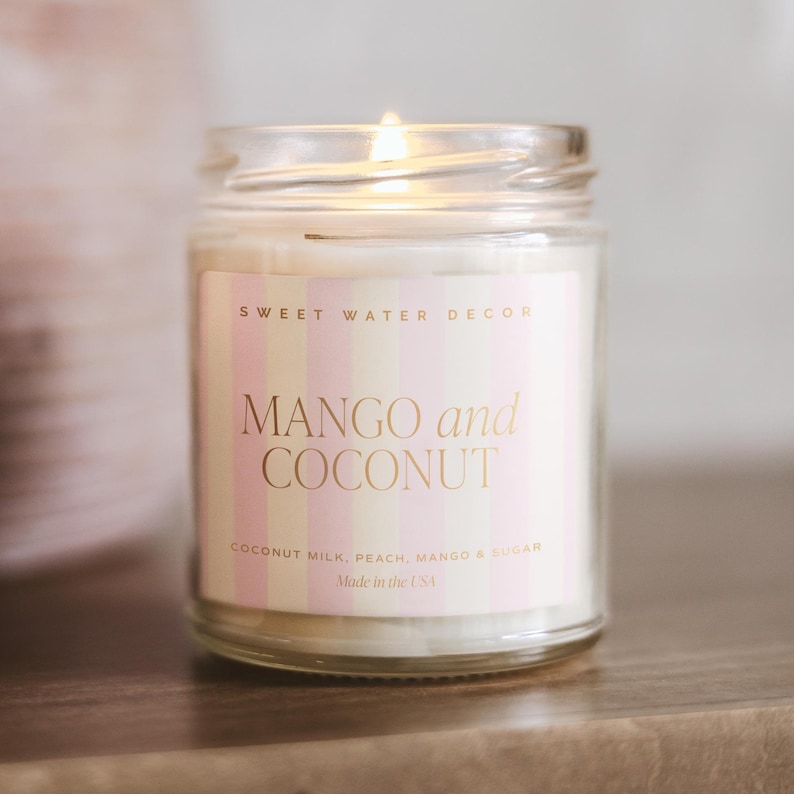 Może przedstawiać: Zapalonego świeca w szklanym słoiku z etykietą z napisem "MANGO and COCONUT". Etykieta ma r&oacute;żowe i białe paski oraz złote litery. Składniki są wymienione pod tytułem. Świeca jest wyprodukowana w USA.