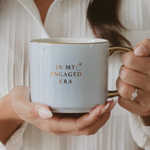 May include: A pale blue ceramic mug with gold detailing. The mug has the text "IN MY ENGAGED ERA" in gold, with a ring graphic. The mug is held by a person.