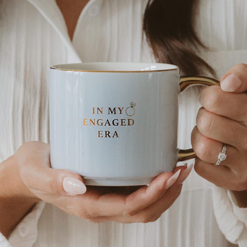 May include: A pale blue ceramic mug with gold detailing. The mug has the text "IN MY ENGAGED ERA" in gold, with a ring graphic. The mug is held by a person.