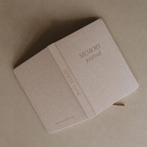 Pu&ograve; includere: Un diario beige rivestito in lino con il testo "MEMORY Journal" in rilievo sulla copertina e sul dorso.