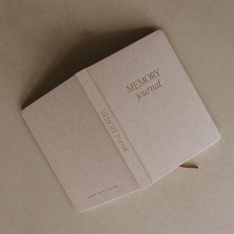 Pu&ograve; includere: Un diario beige rivestito in lino con il testo "MEMORY Journal" in rilievo sulla copertina e sul dorso.