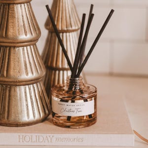 May include: A glass diffuser bottle with black reeds, labeled "Christmas Tree" from Sweet Water Decor, sits on a book with "HOLIDAY memories" printed on it. Gold Christmas tree decorations are in the background.