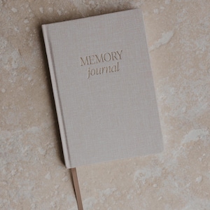 Pu&ograve; includere: Un diario beige rivestito in lino con il testo "MEMORY journal" in rilievo dorato sulla copertina. Il diario ha un segnalibro in nastro marrone.