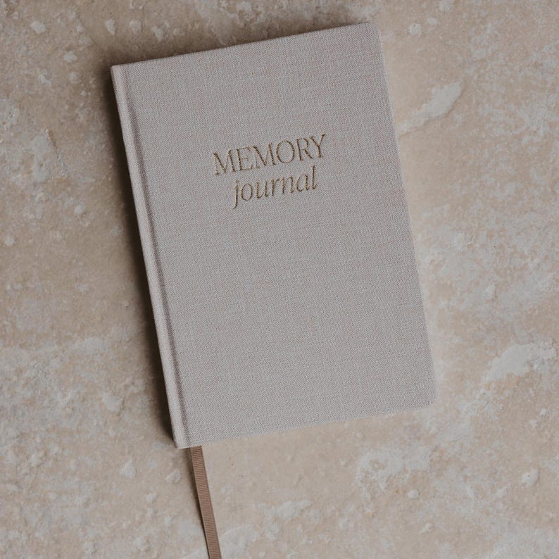 Pu&ograve; includere: Un diario beige rivestito in lino con il testo "MEMORY journal" in rilievo dorato sulla copertina. Il diario ha un segnalibro in nastro marrone.