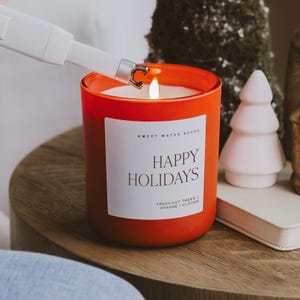 May include: An orange candle in a glass jar is lit with a white lighter. The candle has a white label that reads "HAPPY HOLIDAYS" and "SWEET WATER DECOR". A small white ceramic Christmas tree is next to the candle.