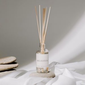 May include: A clear glass bottle with a white label that reads "RELAXATION SWEET WATER DECOR" and wooden diffuser sticks. The bottle is sitting on a light gray stone.