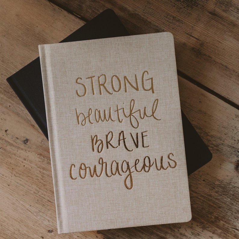 May include: A beige linen journal with the words "STRONG beautiful brave courageous" written in gold lettering.