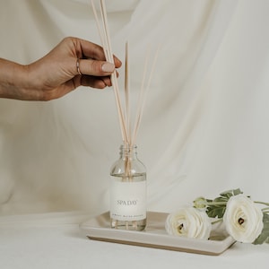 May include: A clear glass aromatherapy diffuser bottle with the text "SPA DAY" and "SWEET WATER DECOR" on a white label. The bottle contains liquid and wooden reeds. The bottle sits on a rectangular, light beige tray, with white flowers.
