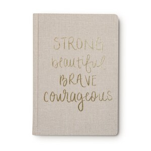 May include: A beige linen-covered journal with the words "STRONG beautiful brave courageous" written in gold lettering.