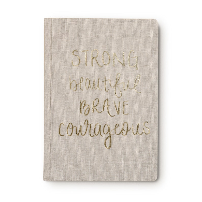May include: A beige linen-covered journal with the words "STRONG beautiful brave courageous" written in gold lettering.