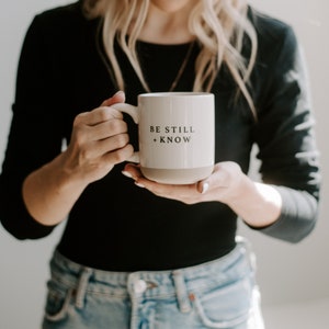 May include: Cream-colored ceramic mug with a light gray base. The mug features the text "BE STILL + KNOW" in black, centered on the front. The mug is held by a person wearing a black long-sleeve shirt and blue jeans.