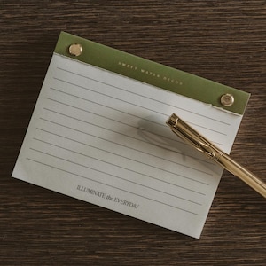 May include: A notepad with lined paper and a green top, secured with gold-colored rivets. The notepad has the text "SWEET WATER DECOR" and "ILLUMINATE the EVERYDAY". A gold pen rests on the notepad.