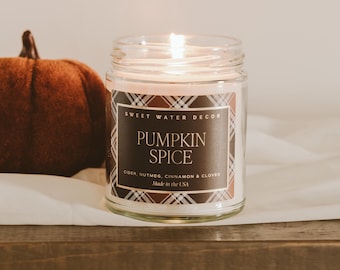 May include: A clear glass jar candle with a lit wick and a label featuring a brown and white plaid border. The label text reads SWEET WATER DECOR, PUMPKIN SPICE, CIDER, NUTMEG, CINNAMON & CLOVES, and Made in the USA. A brown pumpkin sits nearby.