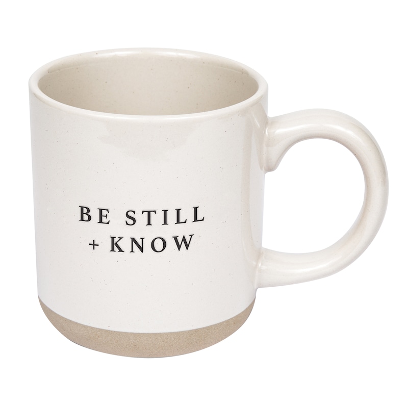 May include: Cream-colored ceramic mug with a rounded handle and a light brown base. The mug features the text "BE STILL + KNOW" in black, centered on the front. The mug has a speckled texture.
