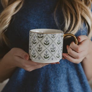 May include: White ceramic mug with a gold handle and rim, featuring a repeating floral pattern in green and light blue. The mug is held by a person wearing a blue top. A decorative and stylish coffee mug.