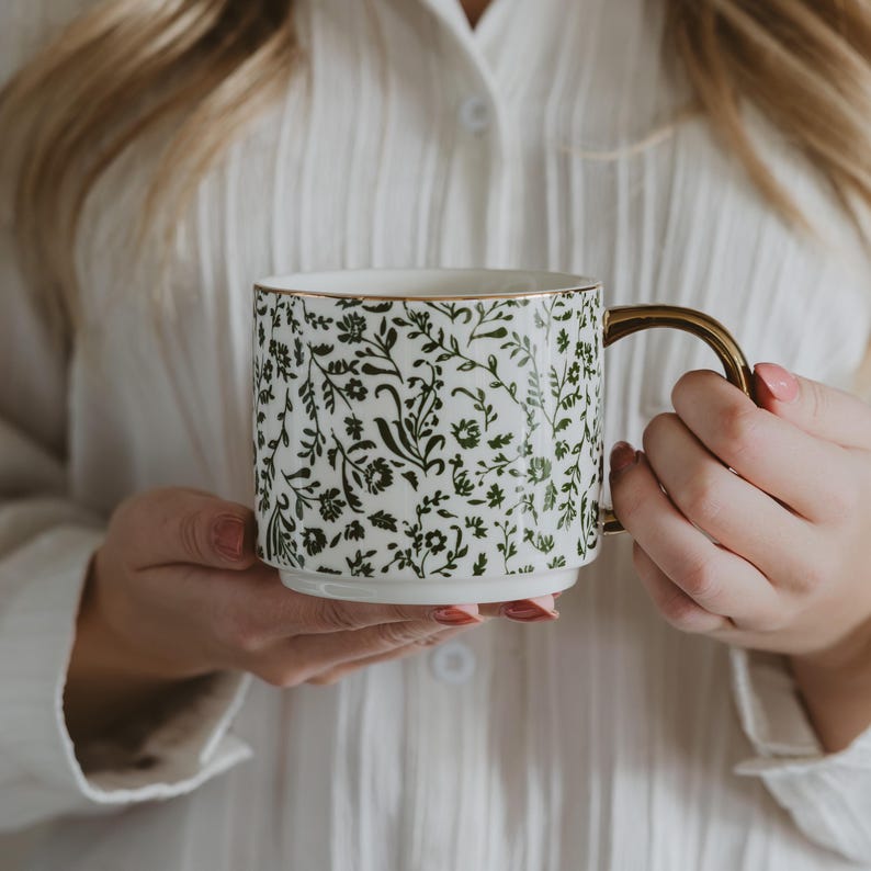 May include: White ceramic mug with a gold handle and rim, adorned with a dark green floral pattern. The mug is held by a person wearing a white shirt. A decorative and stylish coffee mug.