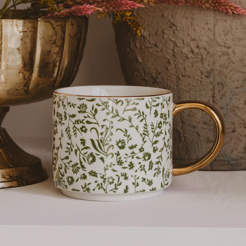 May include: White ceramic mug with a gold handle and rim, adorned with a green floral pattern. The mug is cylindrical and sits on a white surface, with decorative vases in the background.