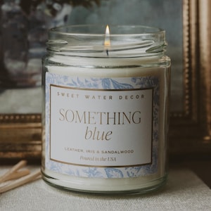 May include: A lit glass jar candle with a white label featuring the text "SOMETHING blue" in gold lettering. The label also includes the scent notes: Leather, Iris & Sandalwood. The candle is in a clear glass jar with a lit wick.