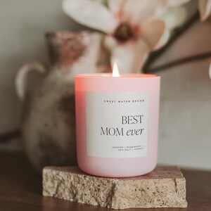 May include: Pink candle with a white label that reads "BEST MOM ever" and "Sweet Water Decor". The candle is on a light brown stone surface. The scent is "Jasmine / Rosemary / Sea Salt / Honey".
