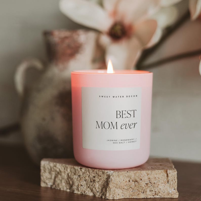 May include: Pink candle with a white label that reads "BEST MOM ever" and "Sweet Water Decor". The candle is on a light brown stone surface. The scent is "Jasmine / Rosemary / Sea Salt / Honey".