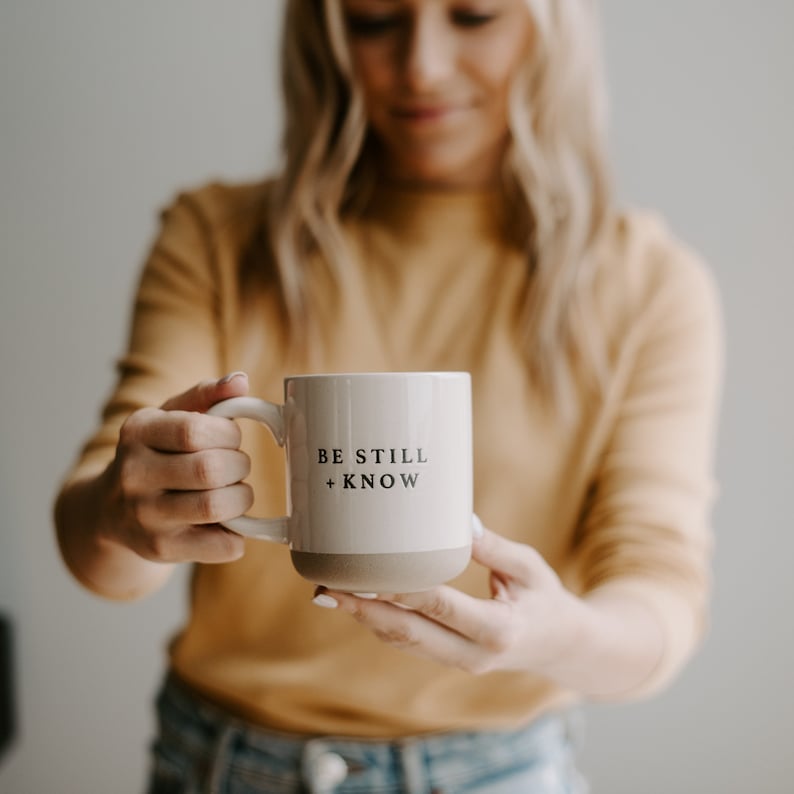 May include: Cream-colored ceramic mug with a light brown base and handle. The mug features the text "BE STILL + KNOW" in black. The mug is held in front of a person wearing a yellow shirt.
