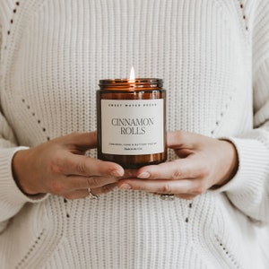 Peut inclure: Une bougie en verre brun avec une &eacute;tiquette blanche qui indique "SWEET WATER DECOR CINNAMON ROLLS CINNAMON, ICING & BUTTERY PASTRY Made in the USA".