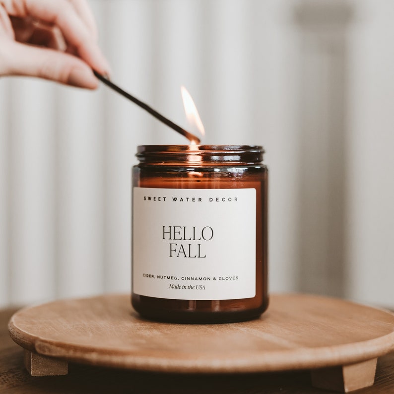 May include: A brown glass jar candle with a white label featuring the text SWEET WATER DECOR, HELLO FALL, CIDER, NUTMEG, CINNAMON & CLOVES, and Made in the USA. The candle has a lit wick with a visible flame, emitting a warm glow from the wax inside.