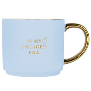 May include: A pale blue ceramic mug with a gold handle and rim. The mug has the gold text "IN MY ENGAGED ERA" and a ring graphic. This mug is designed for hot or cold drinks.