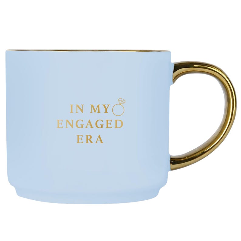 May include: A pale blue ceramic mug with a gold handle and rim. The mug has the gold text "IN MY ENGAGED ERA" and a ring graphic. This mug is designed for hot or cold drinks.