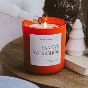 May include: An orange glass candle with a white label that reads "SANTA'S WORKSHOP". The candle is lit with a white lighter. The scent is bayberry, clove, nutmeg, and cedar. A white ceramic Christmas tree is in the background.