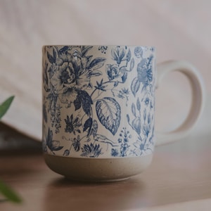 May include: A white ceramic mug with a blue floral pattern. The mug has a wide base and a handle.